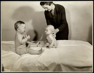 Zwei Babys sitzen im Bett, eines wird von einer Krankenschwester gestützt, das andere hält ein Spielzeugauto und weint im Maternity Center Association an der 70th Street East River, New York, 1935